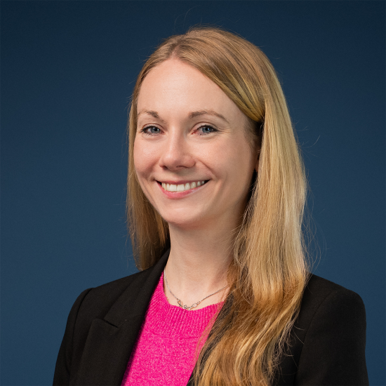 Portrait of Katharina Haen smiling in front of a blue background. She has long blonde hair and is wearing a pink sweater and a black blazer.